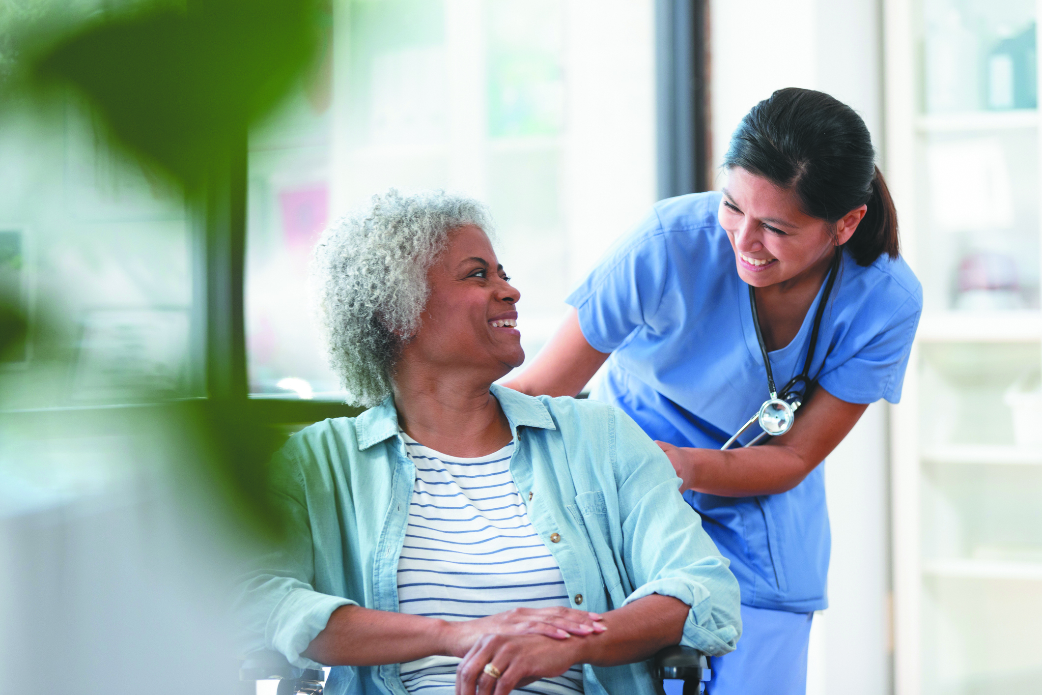 A nurse smiles while pushing a woman's wheelchair into an examination room.