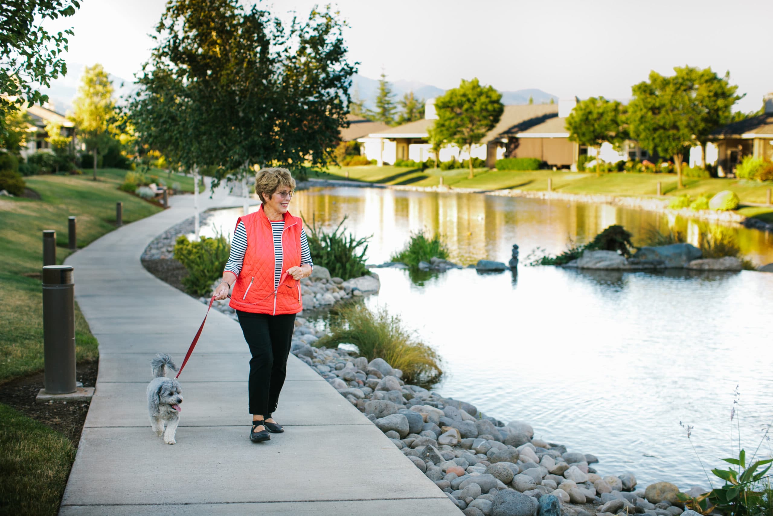 A woman wearing a coral colored vest walks a dog next to a picturesque lake.